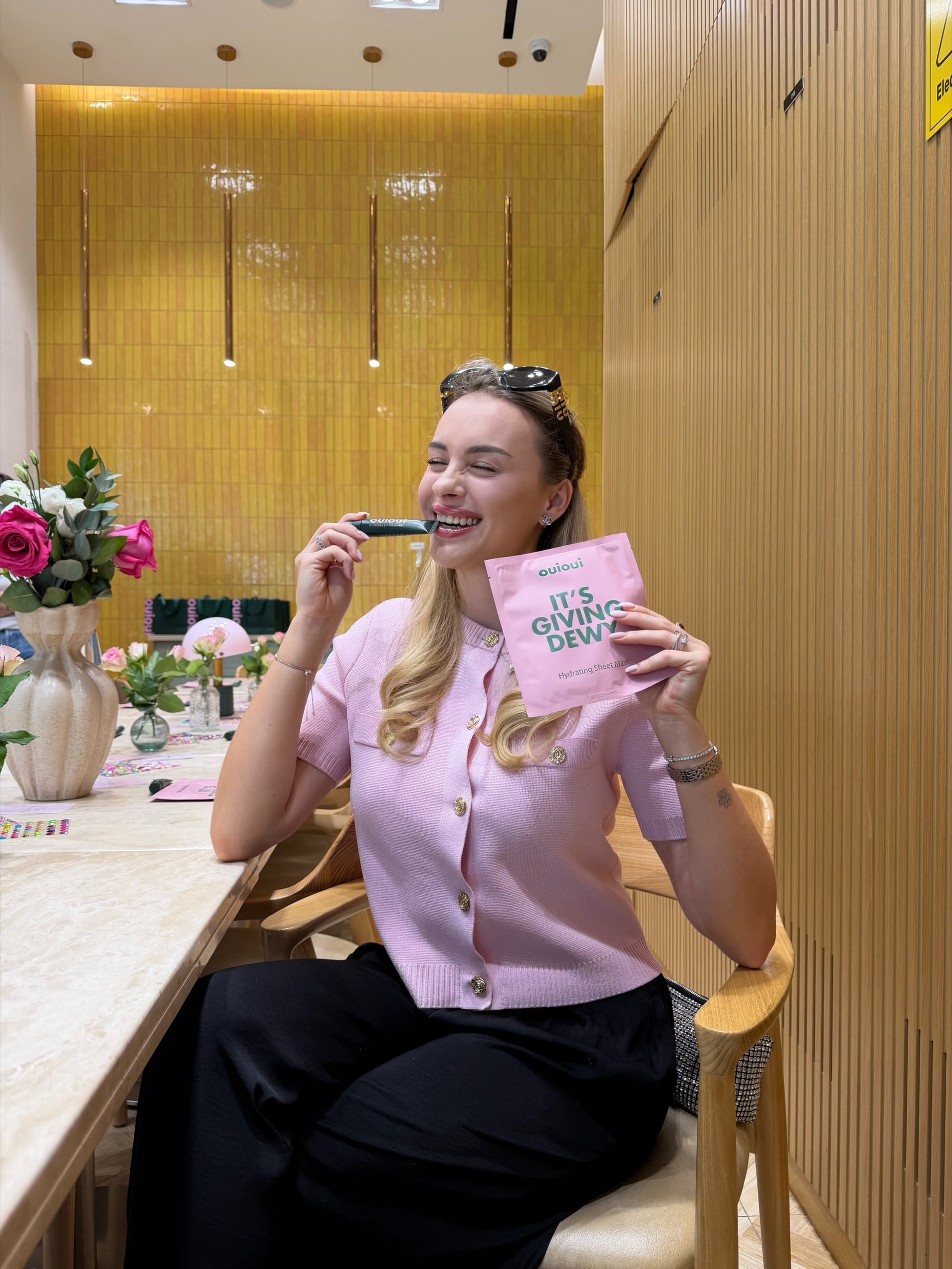 Anna laughing at a café table, holding a pink sheet-mask sachet