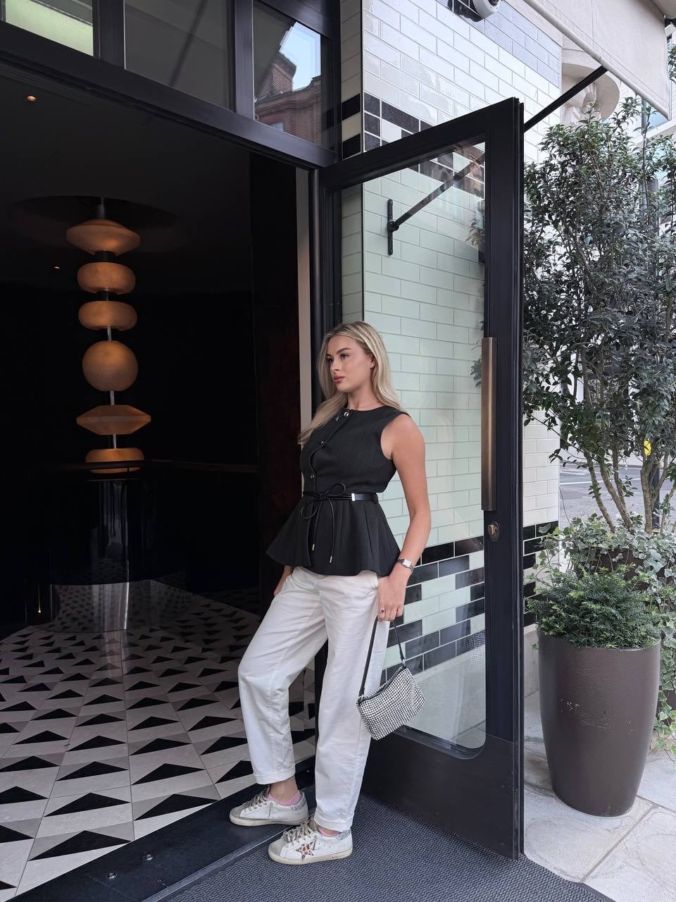 Anna in a black vest and white trousers, leaning in a hotel doorway